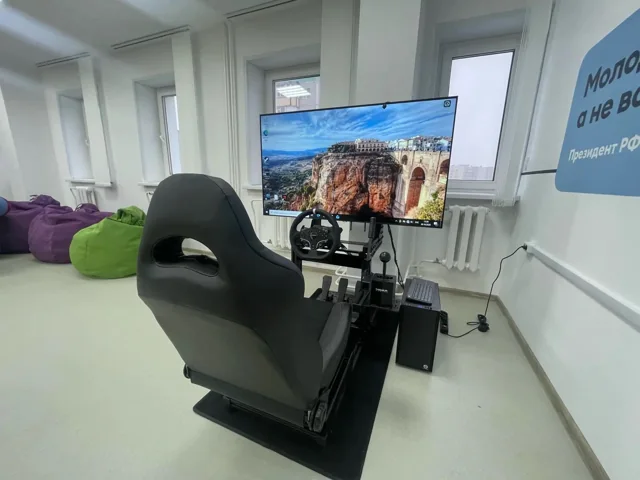 Руль, переключатель передач, педали и компьютер рядом с креслом